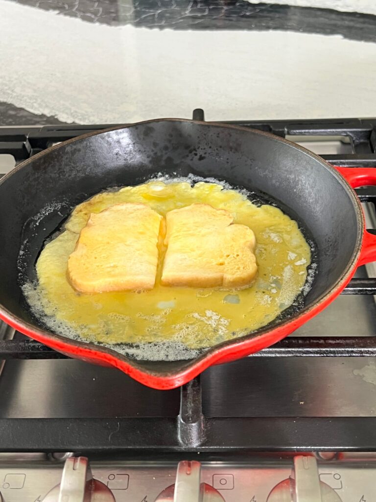 Two bread halves face down in a pan of egg mixture and butter.