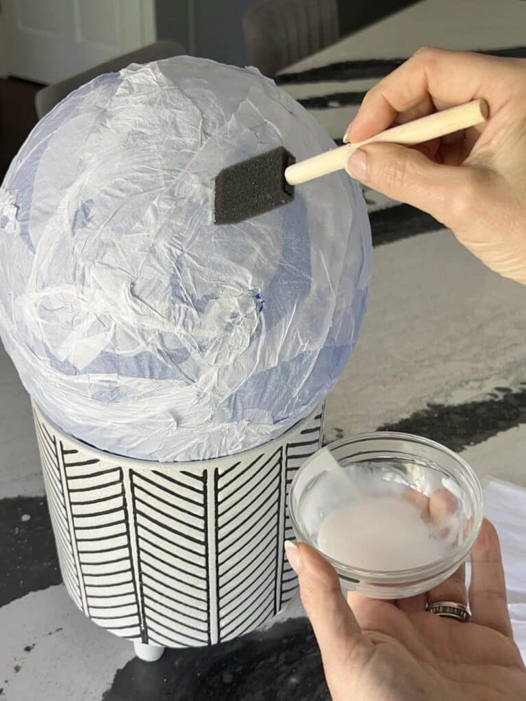 Brushing glue mixture onto tissue covered balloon to create a Diy Tissue Paper Lantern.
