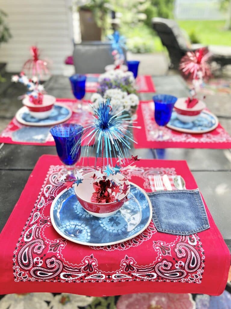 A 4th of July table setting consisting of red bandana placemats, blue plates, red and white bowls, blue goblets, and tinsel firecracker table decor. How to make a placemat.