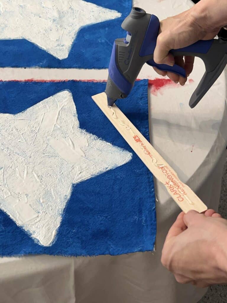 Gluing paint stir sticks to both sides of the blue box on the DIY Flag Banner.