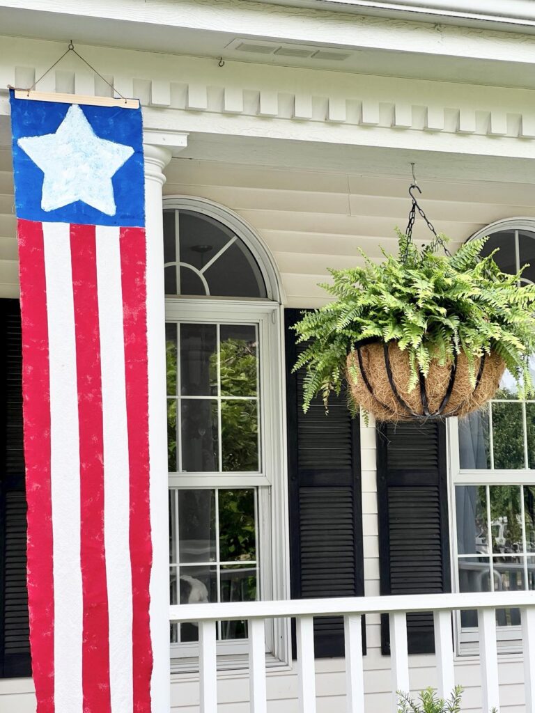 A DIY Flag Banner hanging on a porch beside a hanging fern.