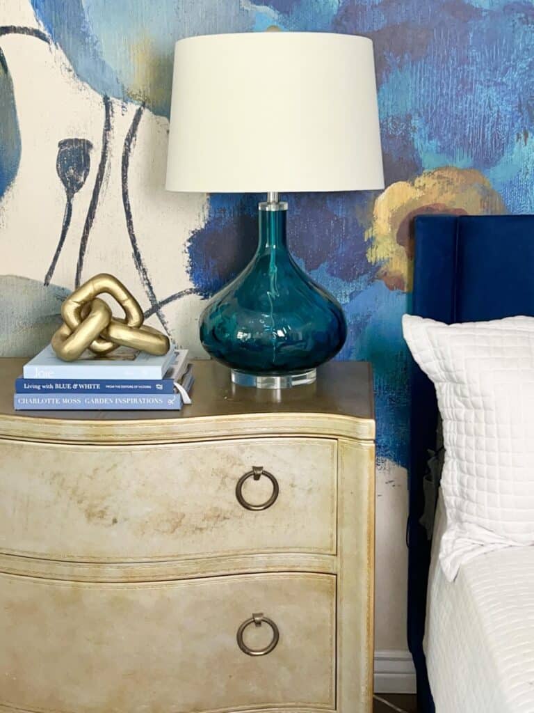 A bedroom nightstand holding a lamp and a stack of blue coffee table books.