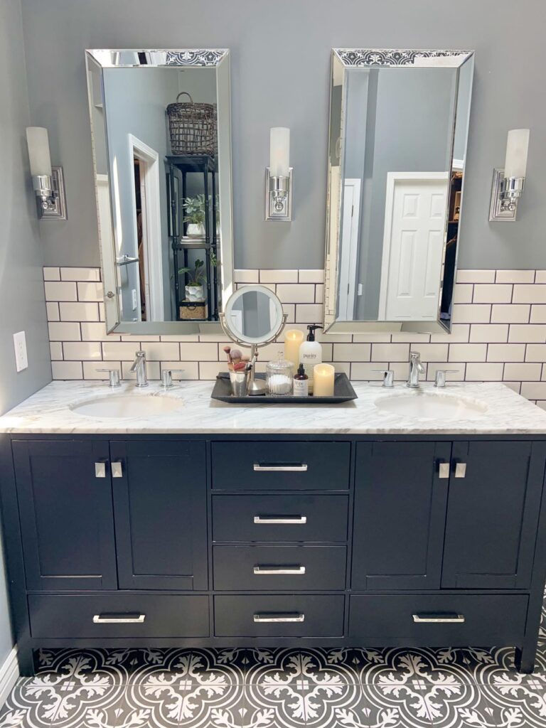 A bathroom counter with a black tray full of home decor ideas like beauty bottles, candles, and bath salts.