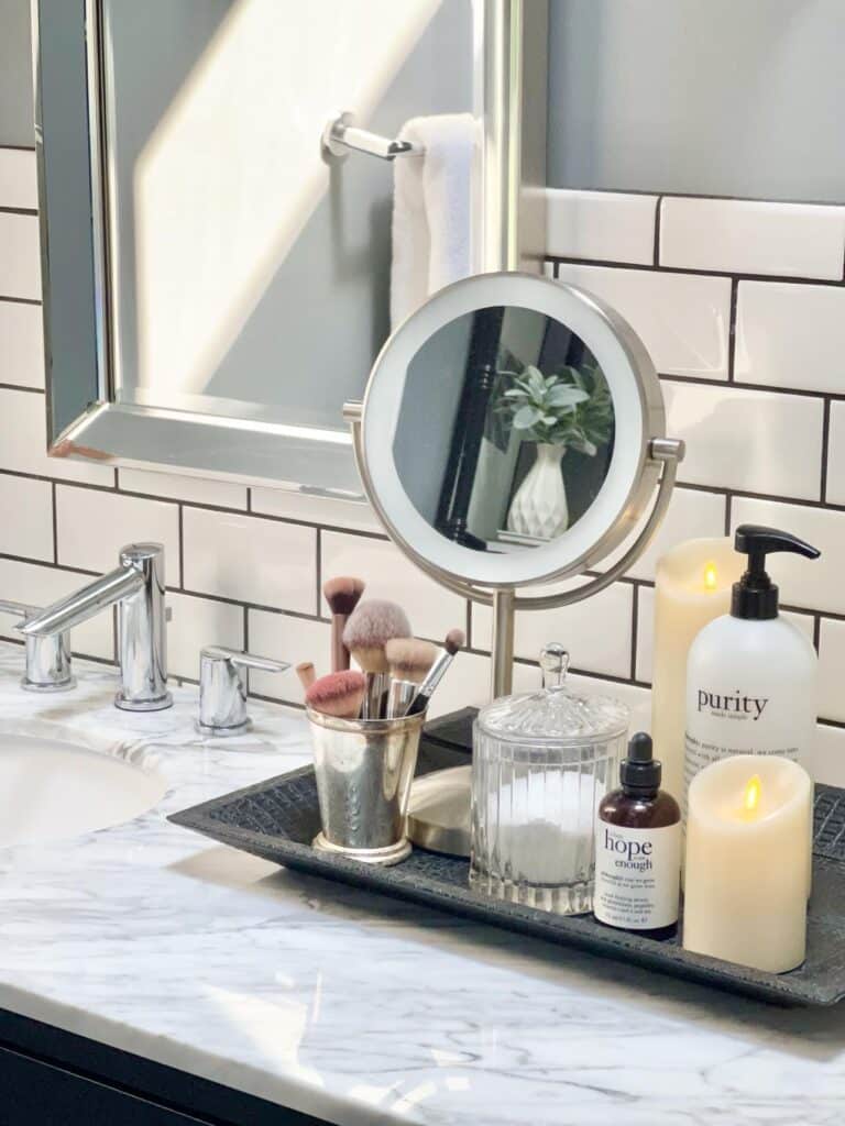 bathroom countertop decor ideas that include a tray with a pedestal mirror, makeup brushes, and faux candles.