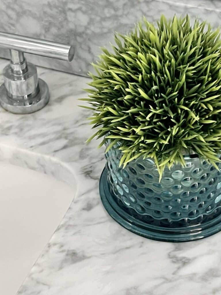 Bathroom countertop decor ideas include a faux green plant in a blue hobnail glass vase.