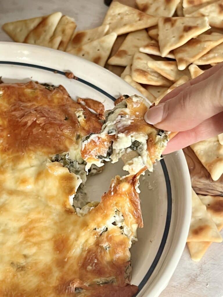 A hand dipping a pita chip into baked spinach artichoke dip.