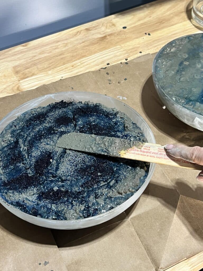 Smoothing blue colored concrete mix into glass bowl with a paint stir stick.