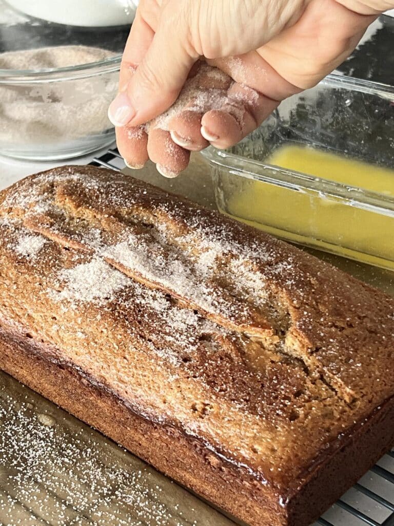 Sprinkling cinnamon sugar to the top of a loaf of apple cider donut bread.