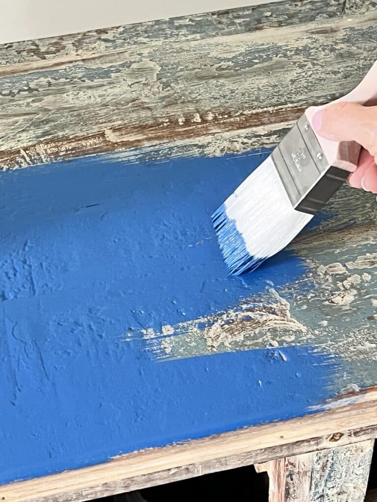 Brushing blue paint onto a furniture piece with a paintbrush for a smooth painted finish.