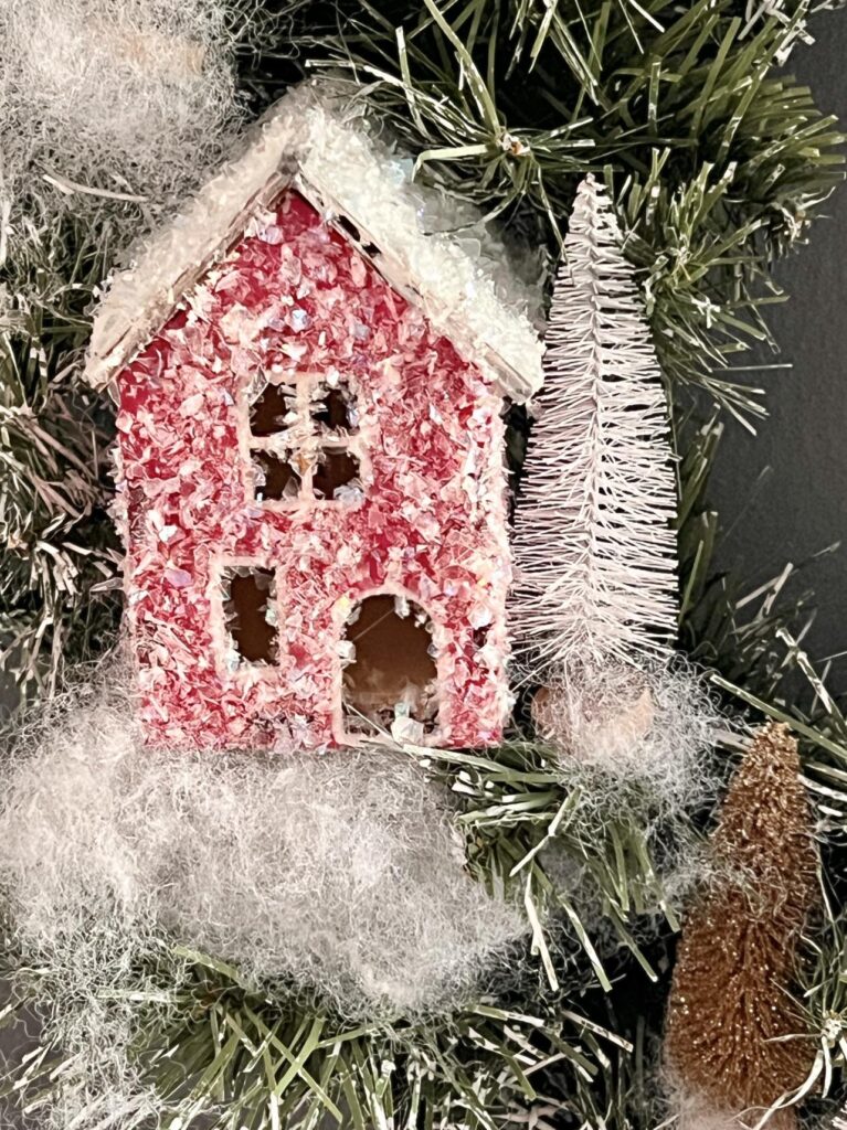 A red snow glittered house nestled into an Anthropologie DIY Christmas Wreath with a white bottle brush tree.