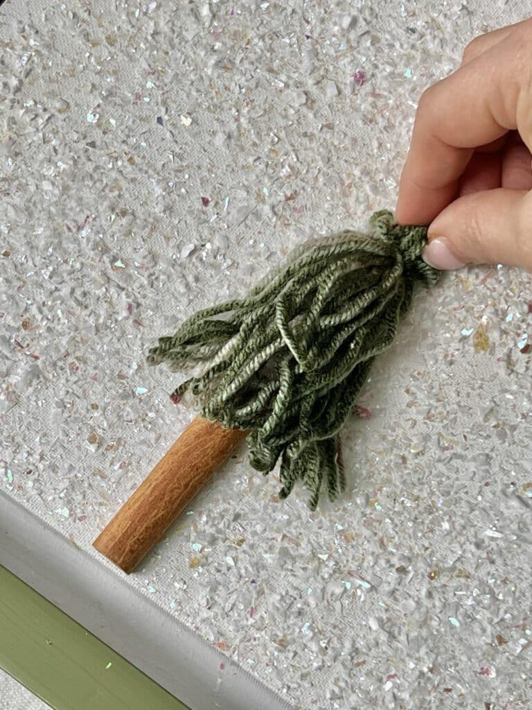 Gluing a Christmas tree tassel over the top end of a cinnamon stick glued to a canvas.