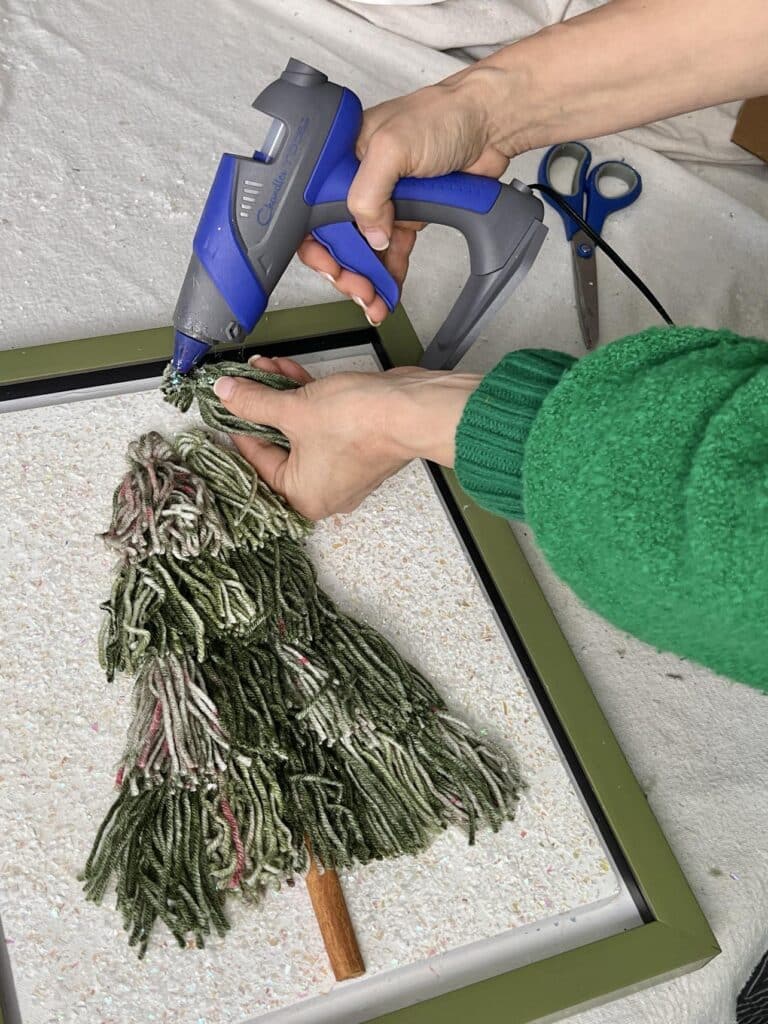 Hot gluing Christmas tree tassels in a pyramid shape on a canvas.