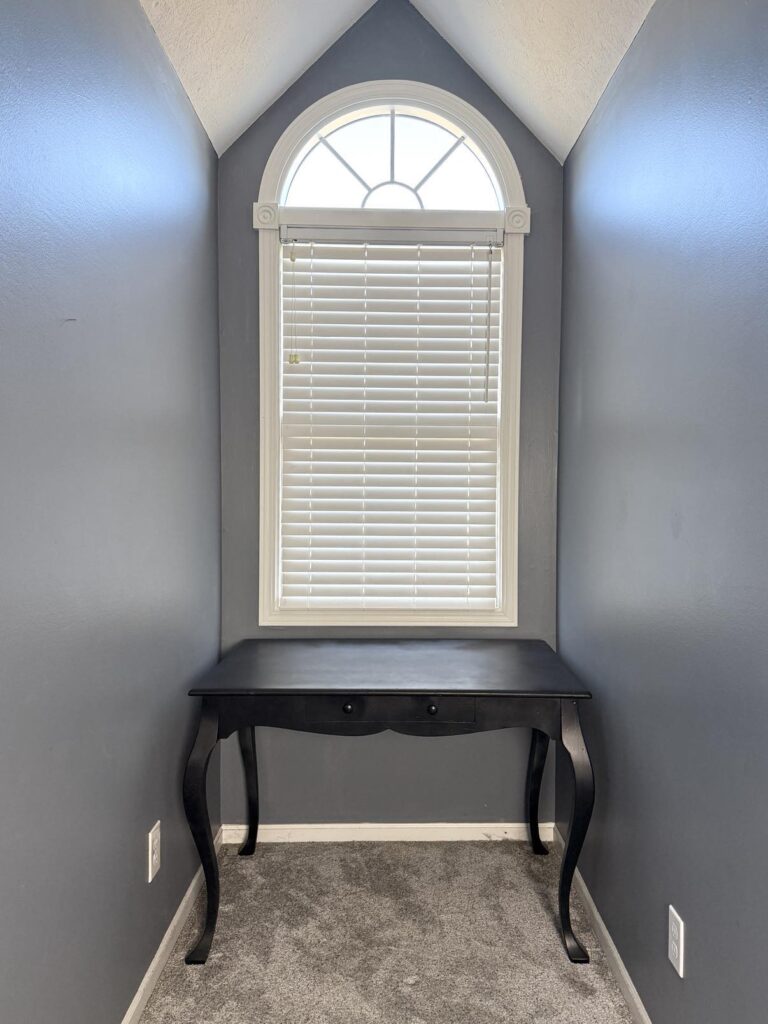 A black table desk in a bedroom nook with a tall window.