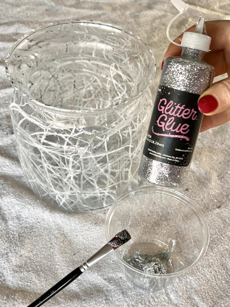 A bottle of silver glitter glue beside a glass vase with crayon markings on the outside.