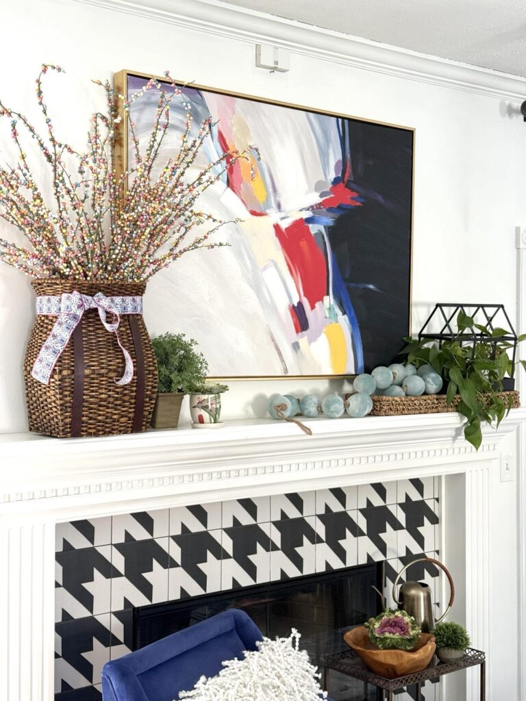 A fireplace mantel decorated for spring with bold artwork, baskets, plants, and bead garland.